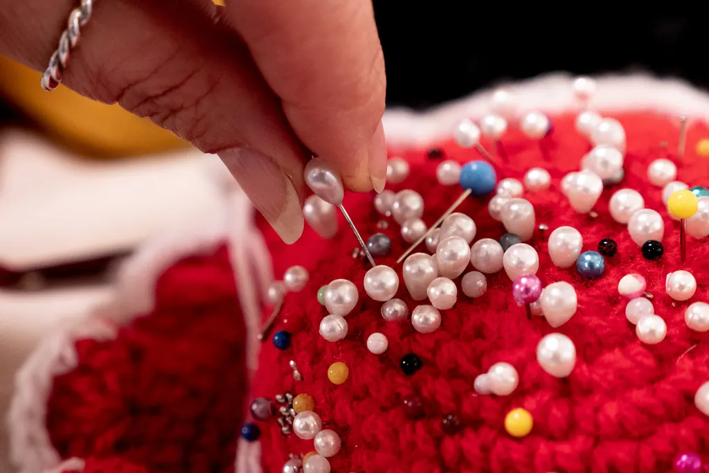 A hand placing a pearl-headed pin into a red pin cushion